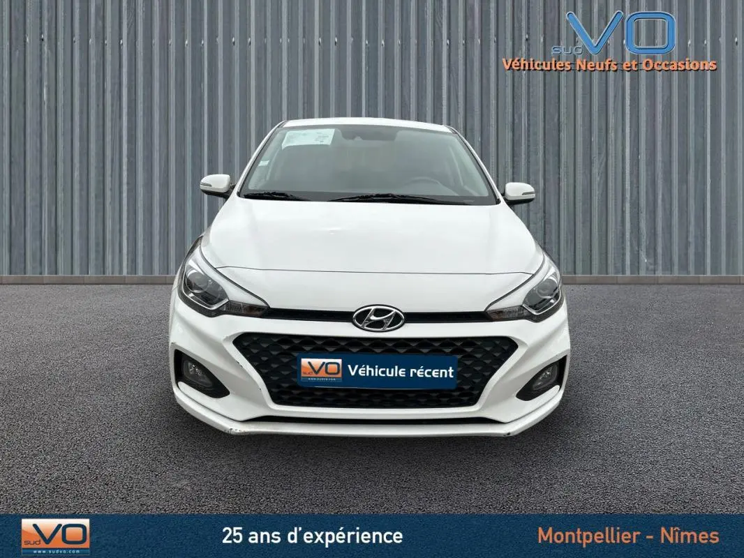 Vue frontale d'une Hyundai i20 blanche de 2019 avec calandre noire et phares LED allumés.