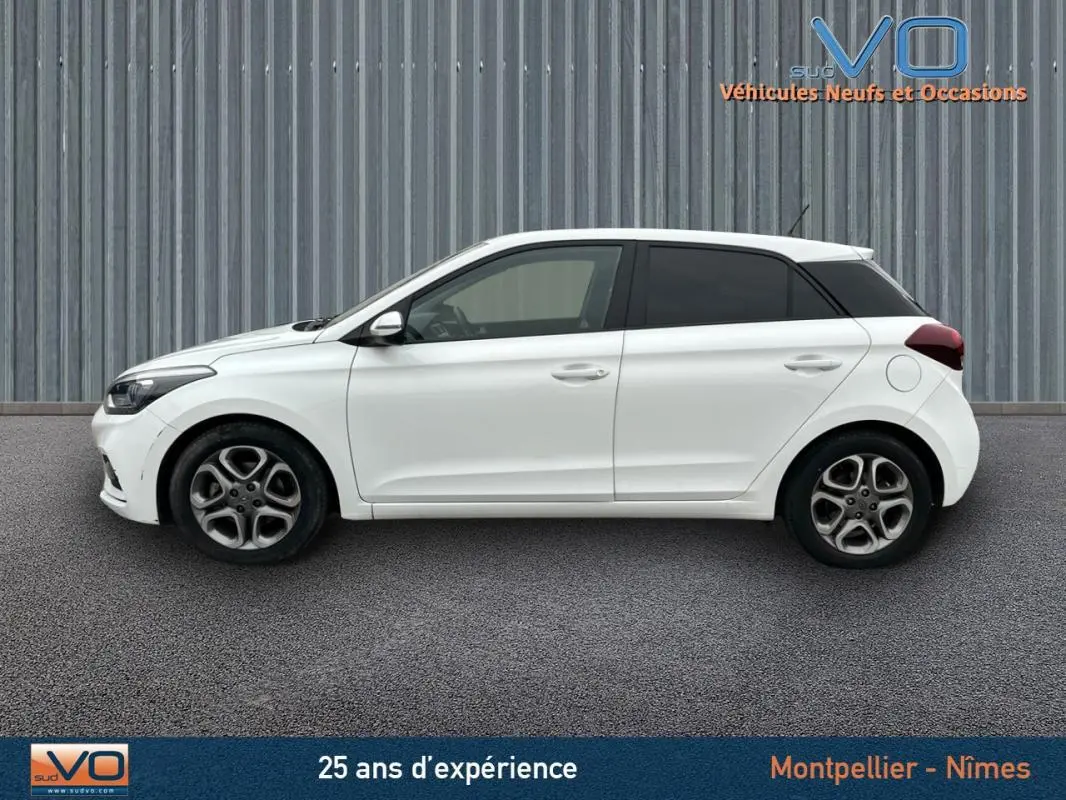 Profil côté gauche d'une Hyundai i20 blanche de 2019 avec jantes alliage et vitres teintées sur fond industriel.