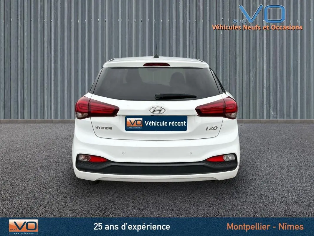 Vue arrière d'une Hyundai i20 blanche de 2019 avec feux rouges et lunette arrière teintée sur fond industriel.