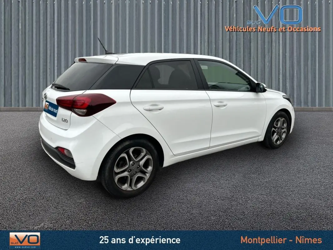 Vue 3/4 arrière droite d'une Hyundai i20 blanche de 2019 avec jantes alliage et vitres teintées arrière.