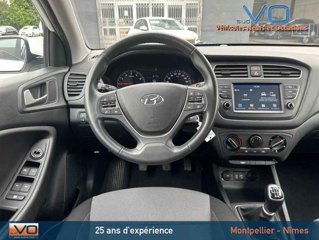 Vue intérieure frontale du tableau de bord et volant d'une Hyundai i20 blanche 2019 avec boîte manuelle et écran tactile central.
