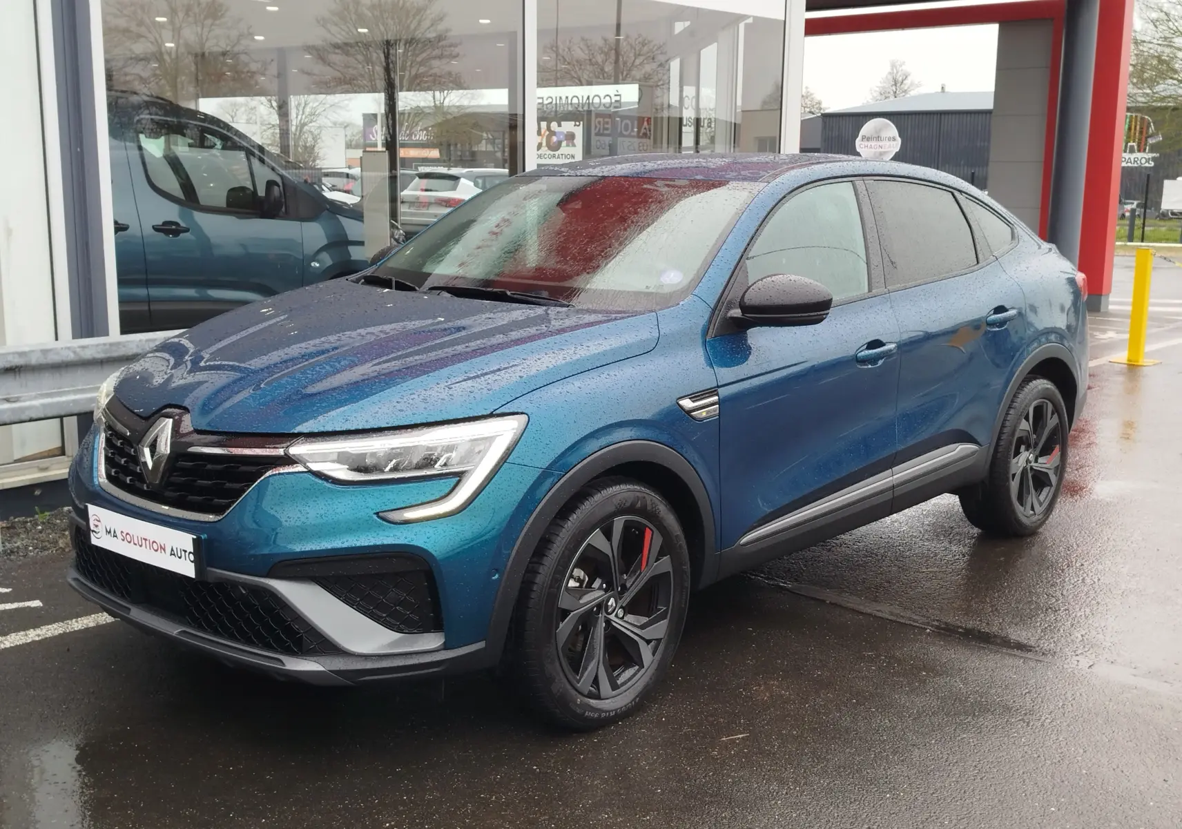 Renault Arkana 1.6 E-Tech bleu Zanzibar en 3/4 avant droit, avec jantes noires et détails rouges sous la pluie.