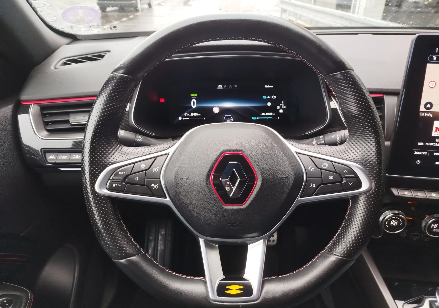Vue intérieure centrée sur le volant noir perforé avec surpiqûres rouges du Renault Arkana bleu Zanzibar, tableau de bord digital visible.