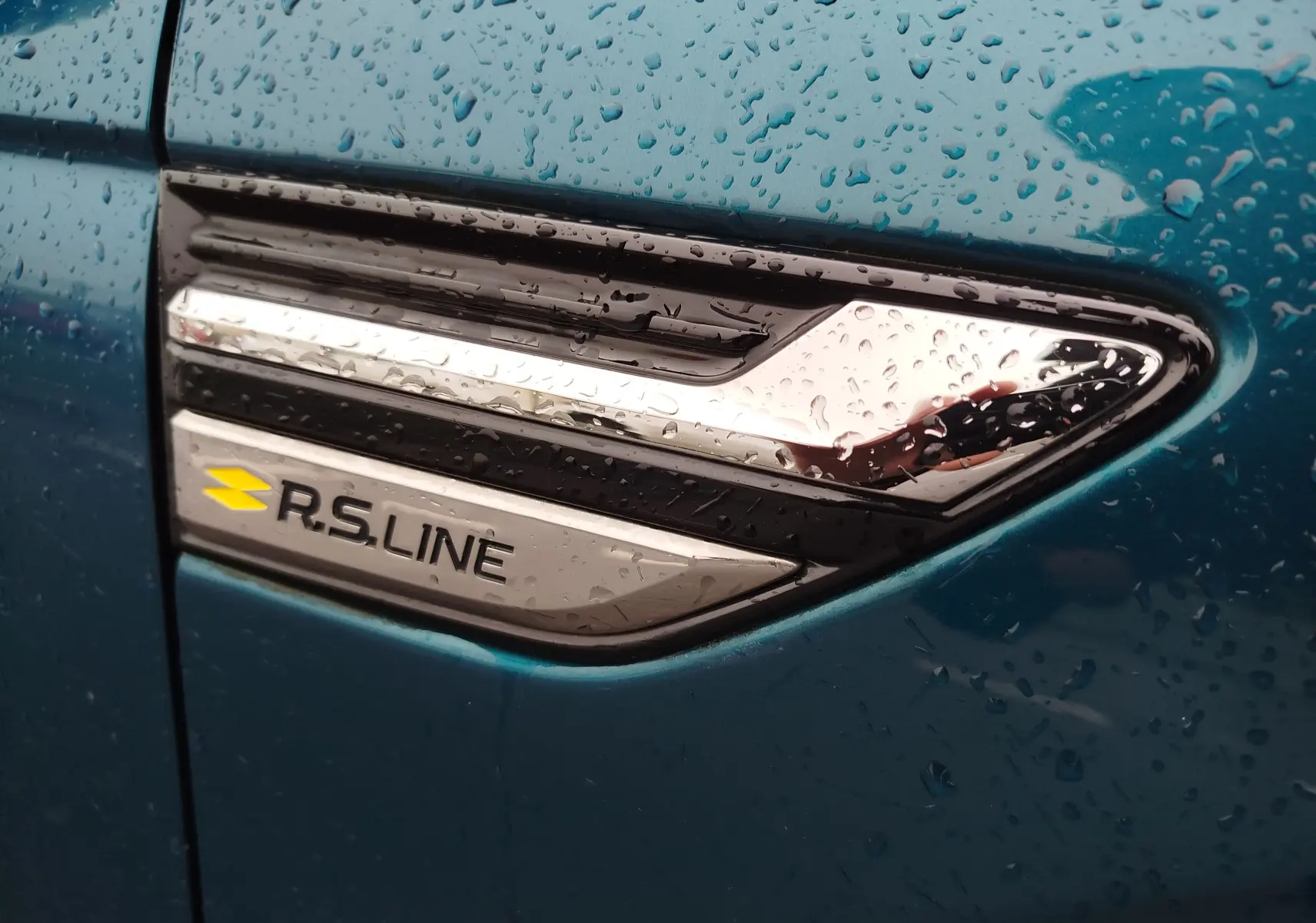 Gros plan sur l'emblème R.S. Line chromé sur la portière bleu Zanzibar d'une Renault Arkana 2022, avec gouttes de pluie.