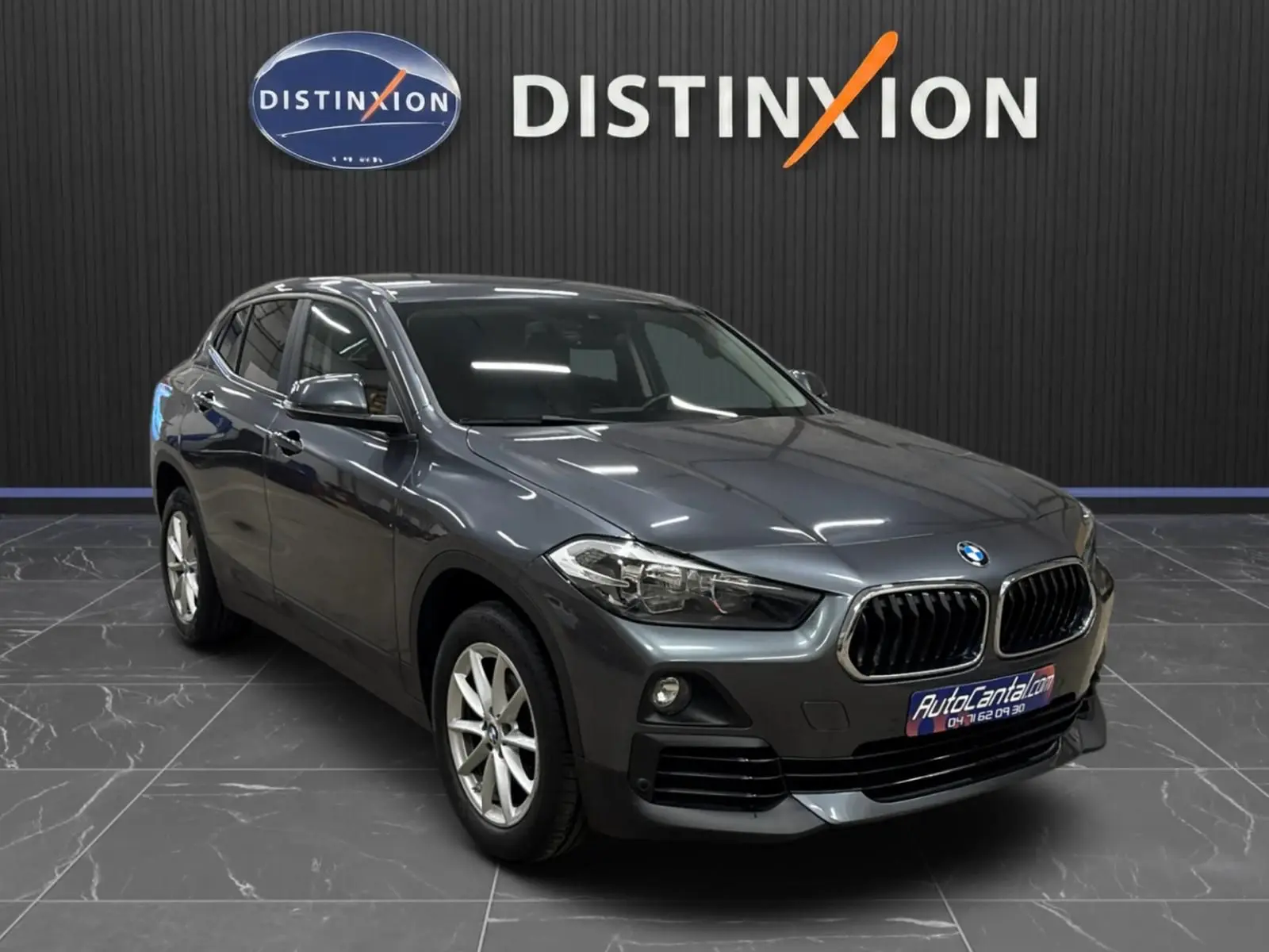 BMW X2 gris minéral métallisé vue 3/4 avant droit dans un showroom avec logo Distinxion en arrière-plan.