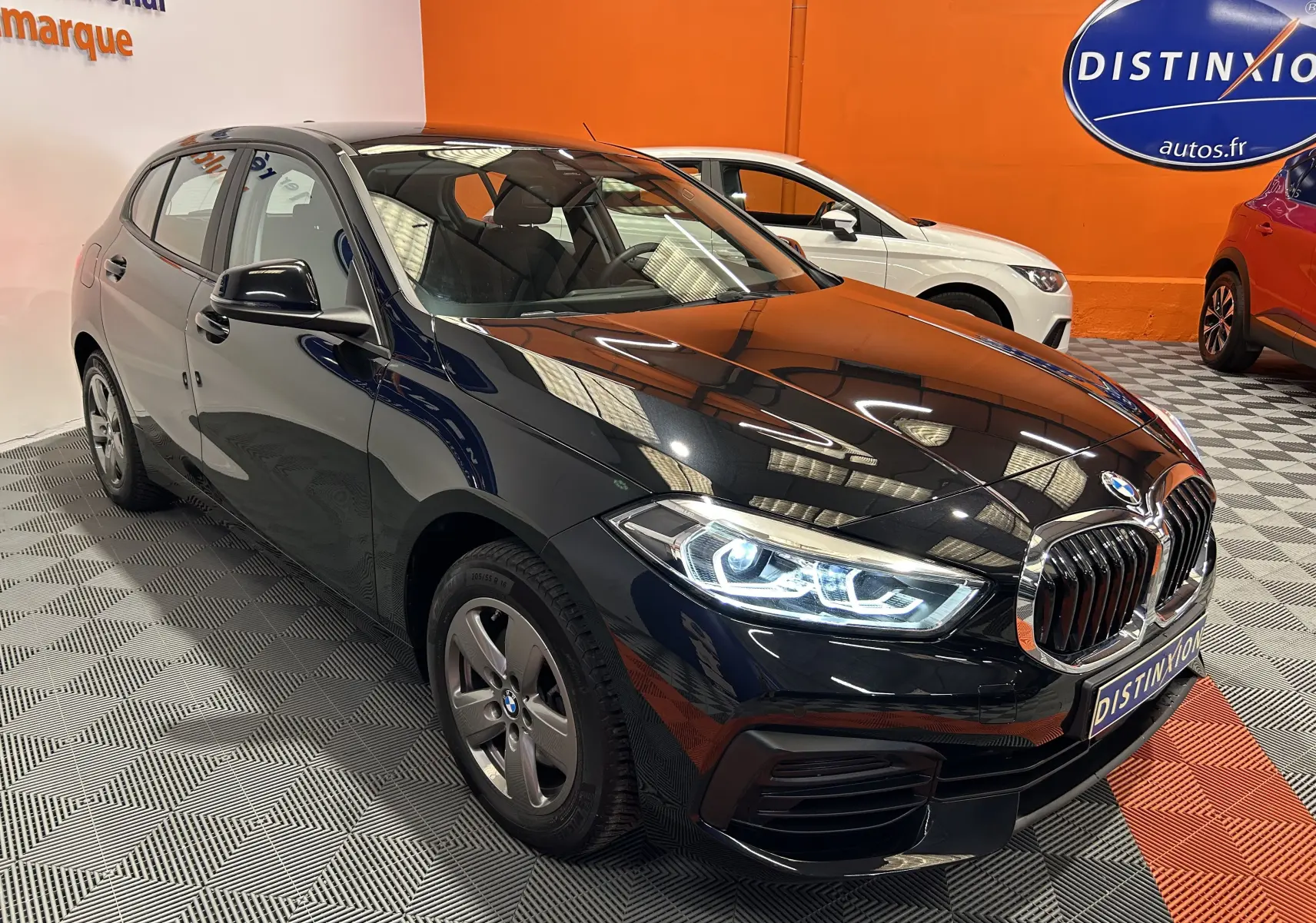 BMW Série 1 16d noire vue en 3/4 avant droit, phares allumés, dans un showroom avec sol gris et orange.