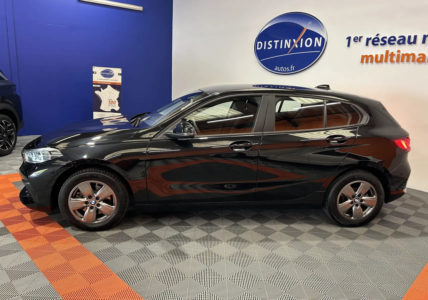 Profil côté gauche d'une BMW Série 1 noire 5 portes, exposée en intérieur avec un sol à motifs gris et orange.