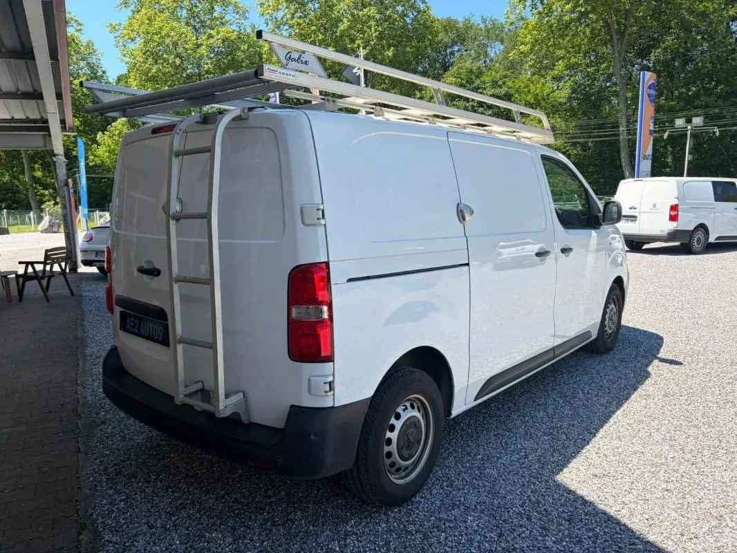Vue 3/4 arrière droite du Peugeot Expert Fourgon blanc avec galerie et échelle sur le toit, stationné sur gravier.