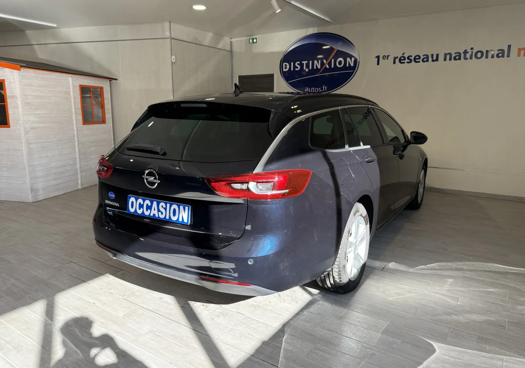 Vue 3/4 arrière droite d'une Opel Insignia Sports Tourer bleu lune marine métal en intérieur avec plaque "OCCASION" visible.