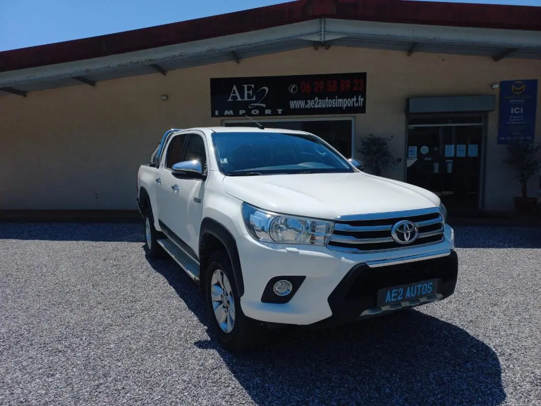 Toyota Hilux 4x4 blanc en 3/4 avant droit, double cabine, avec calandre chromée et barres de toit visibles.