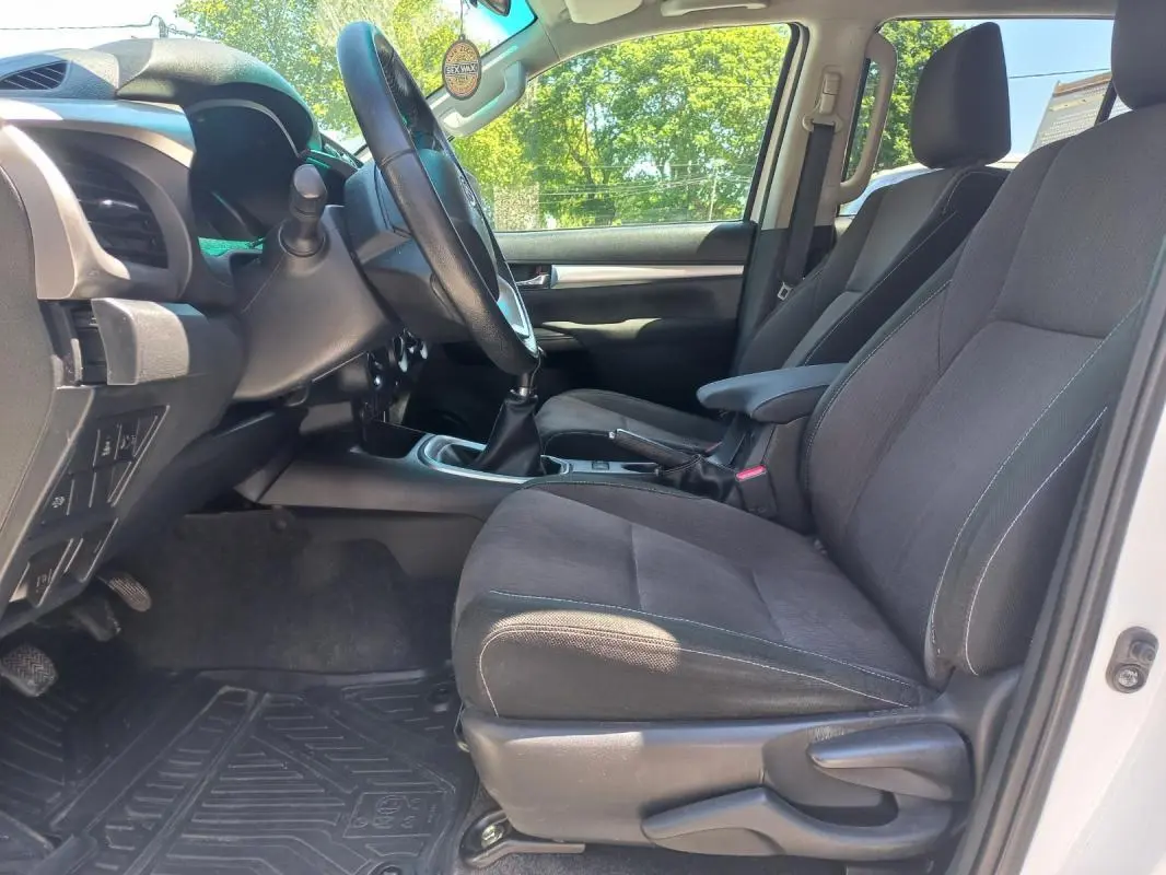 Vue intérieure côté conducteur du Toyota Hilux blanc 2018, montrant les sièges noirs et la boîte manuelle.