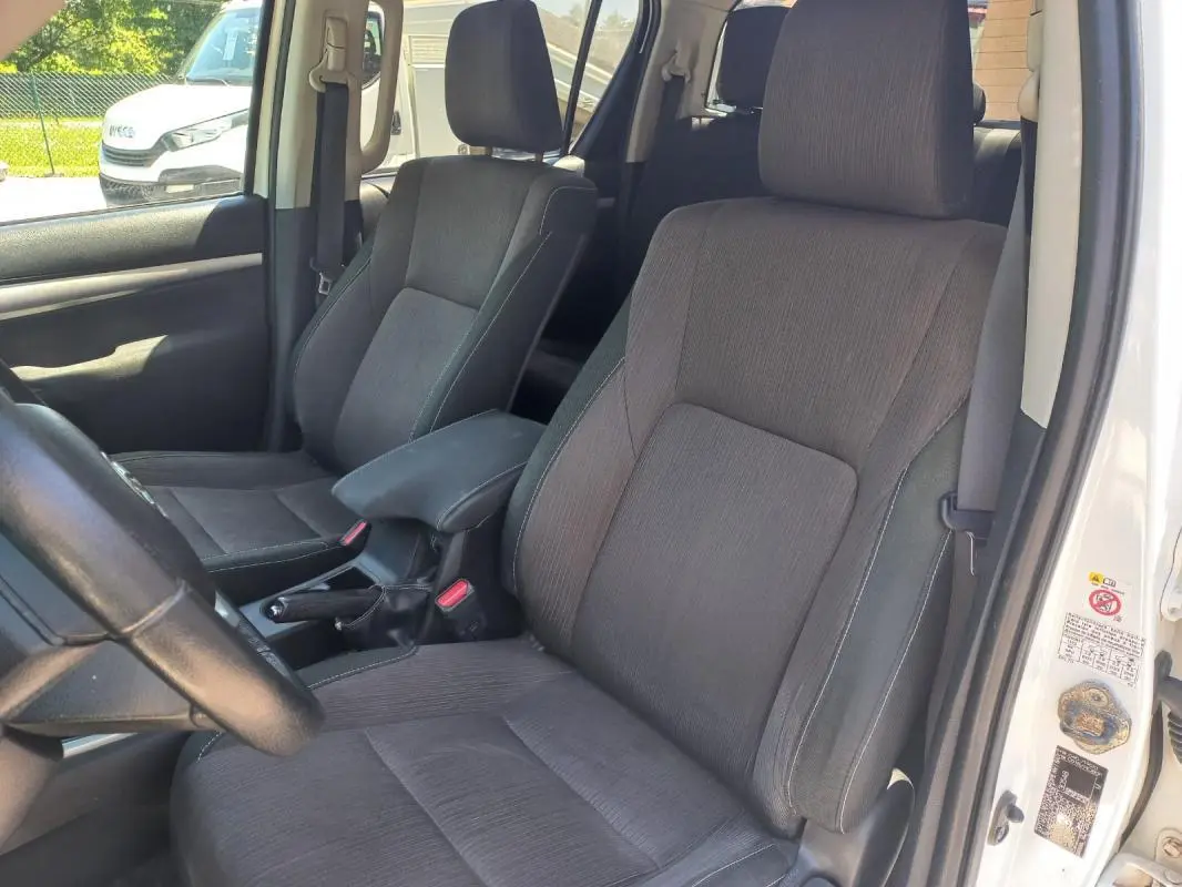 Vue intérieure côté conducteur du Toyota Hilux blanc 2018, montrant les sièges en tissu gris et la boîte manuelle.