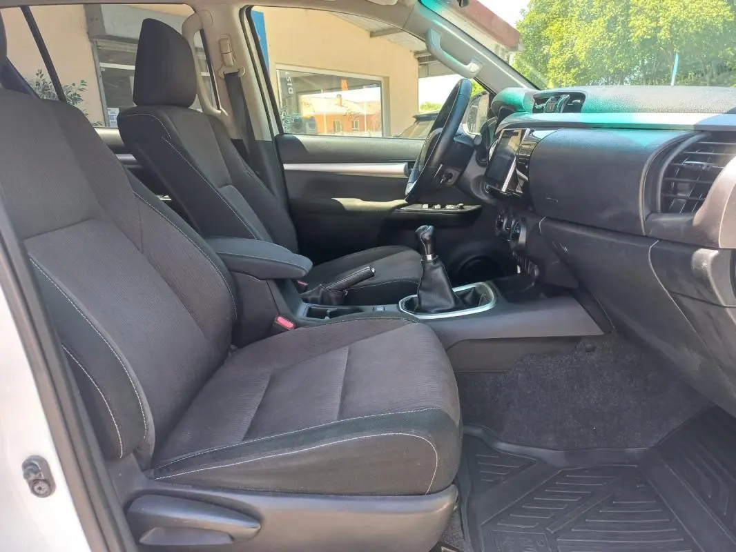 Vue intérieure côté conducteur du Toyota Hilux blanc 2018, mettant en valeur les sièges noirs et la boîte manuelle.