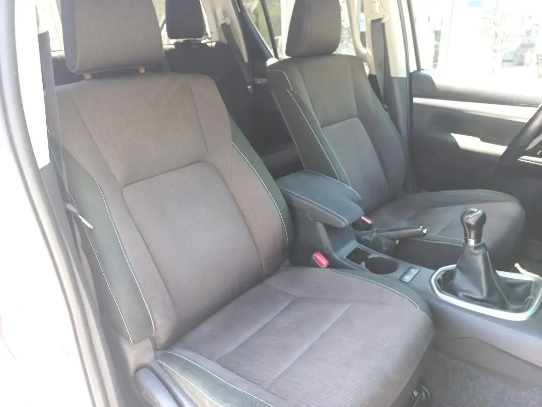 Vue intérieure côté conducteur du Toyota Hilux blanc 2018, montrant les sièges en tissu gris et la boîte manuelle.