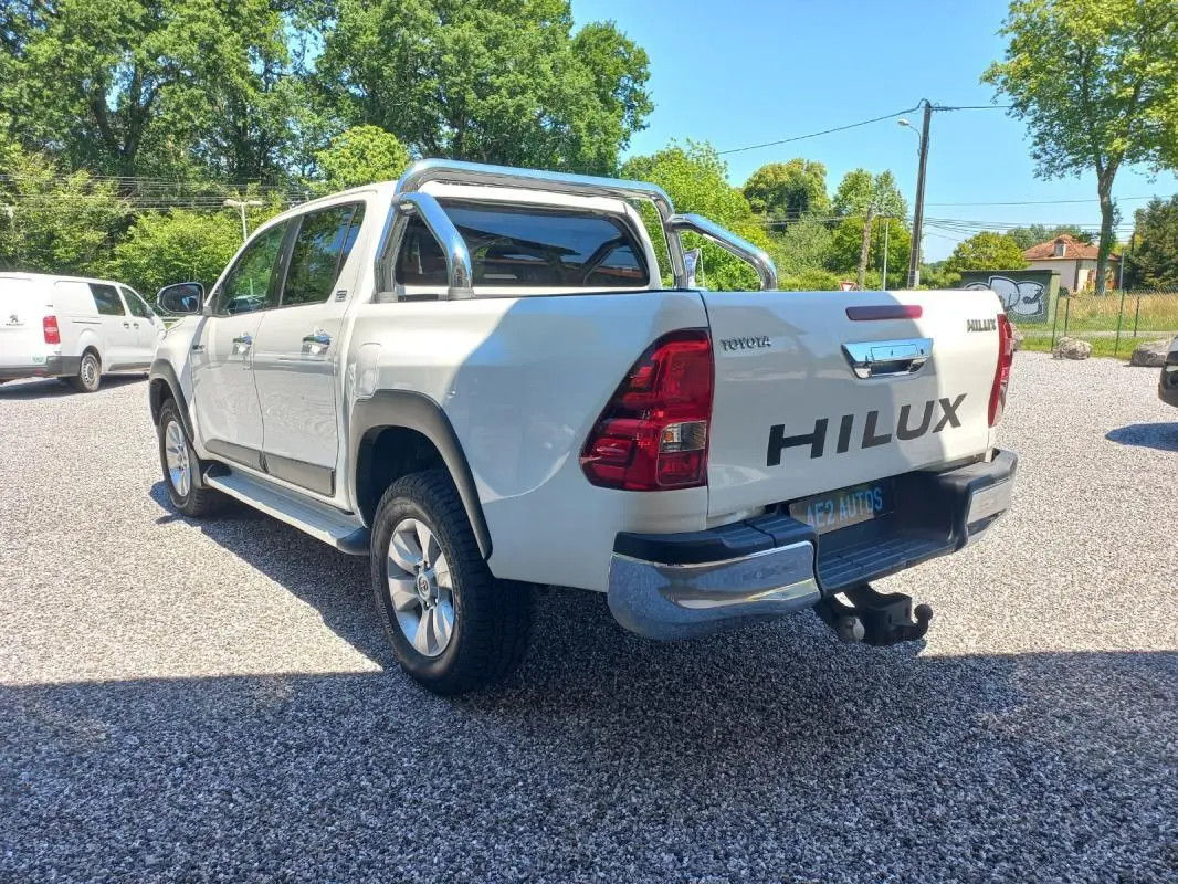 Toyota Hilux 2018 blanc en 3/4 arrière droit, double cabine avec barres chromées et porte-remorque visible.