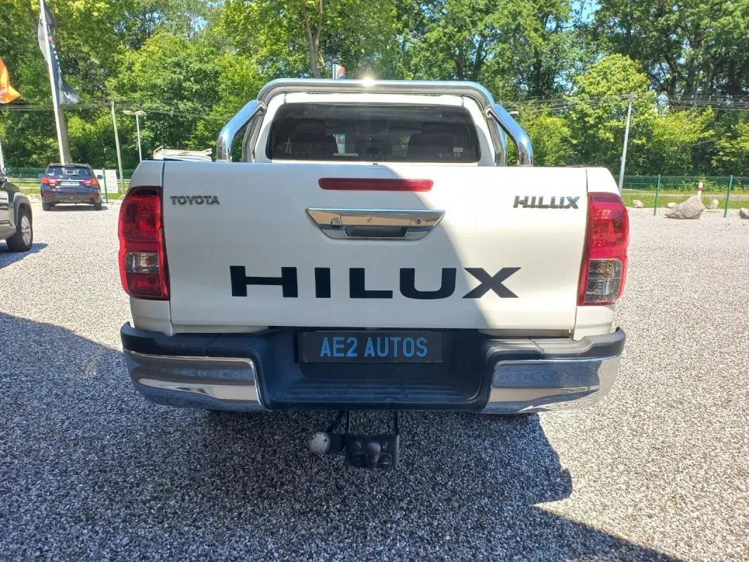 Vue arrière d'un Toyota Hilux blanc 4x4 avec hayon marqué HILUX et attelage visible, stationné en extérieur.