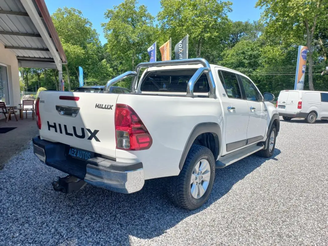 Toyota Hilux blanc en 3/4 arrière droit, double cabine avec barre de protection chromée sur benne, pneus tout-terrain visibles.