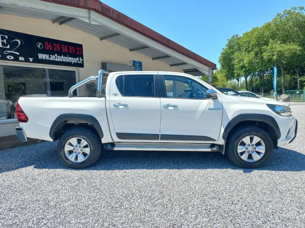 Profil côté gauche du Toyota Hilux 4x4 blanc double cabine avec barre de protection chromée, roues tout-terrain visibles.