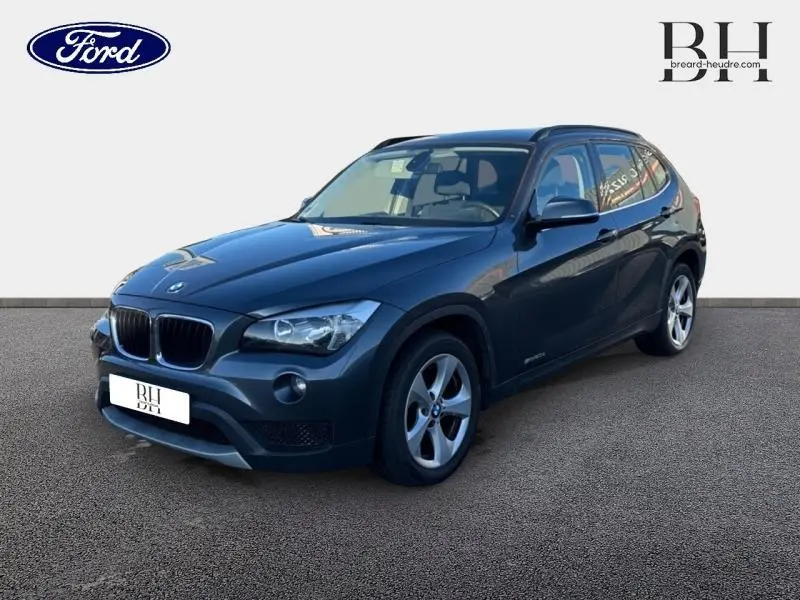 BMW X1 sDrive20d 2013 Mineralgrau en 3/4 avant droit avec jantes alu 17 pouces et barres de toit noires mat.