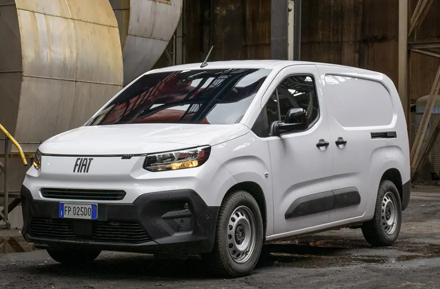 Fourgon Fiat Doblo blanc en 3/4 avant droit, avec calandre noire et feux allumés dans un environnement industriel.
