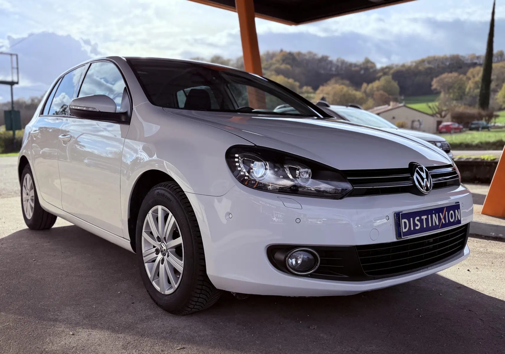 Volkswagen Golf blanche vue en 3/4 avant droit sous abri avec phares allumés et jantes argentées.
