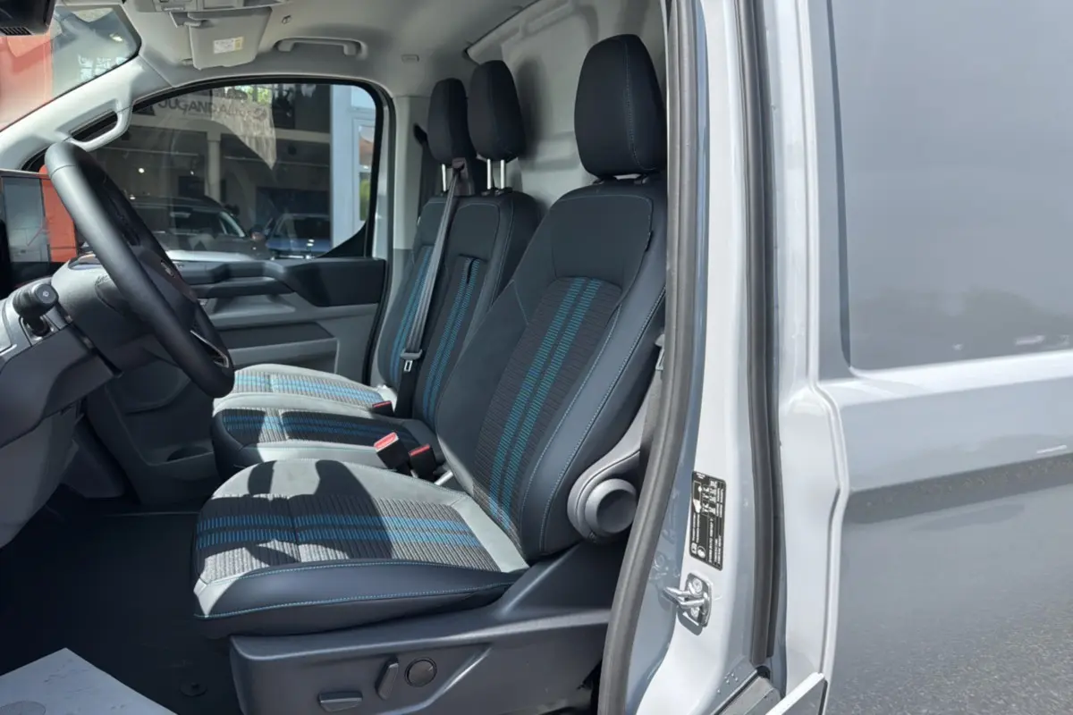 Vue intérieure côté conducteur du Ford Transit Fourgon Custom gris matter, montrant les trois sièges avant en tissu avec détails bleus.