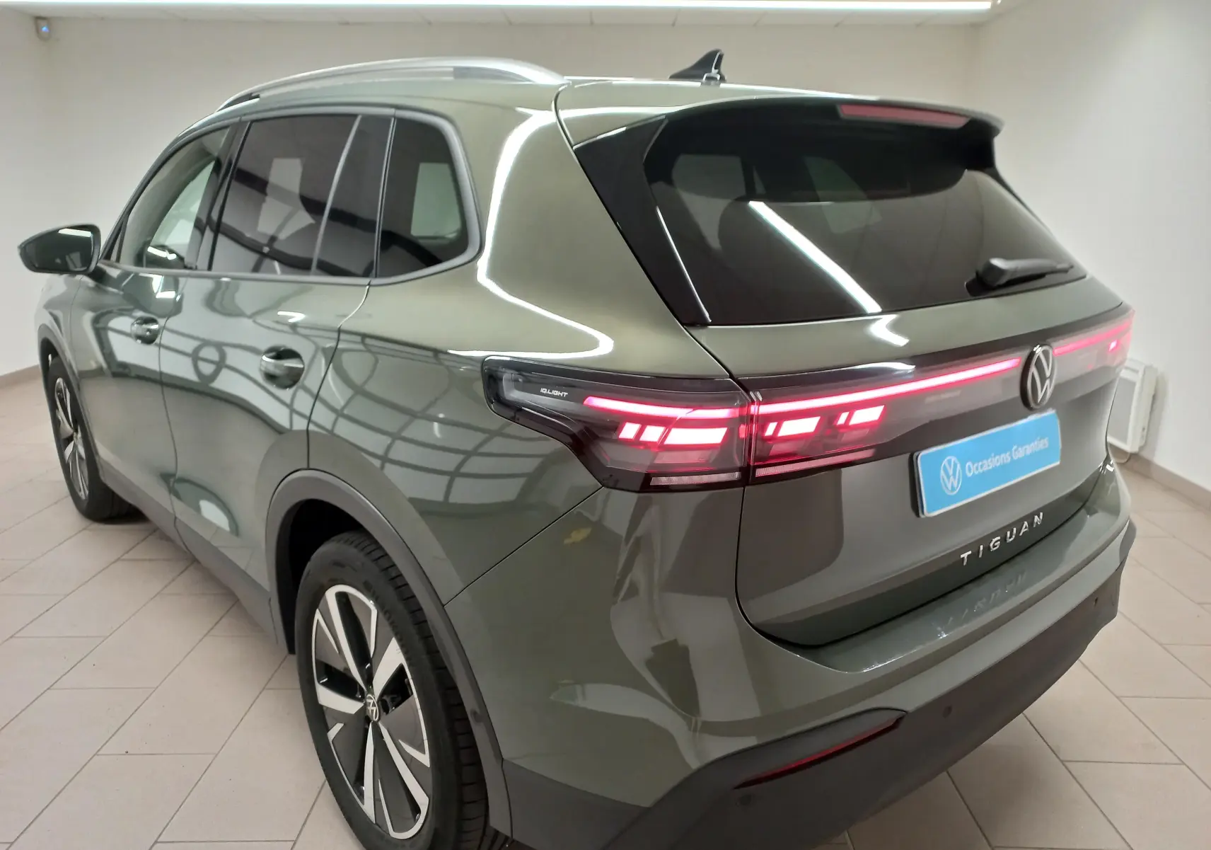 Vue 3/4 arrière gauche d'un Volkswagen Tiguan 2025 vert Cipressino avec jantes alliage noir brillant et feux arrière allumés.