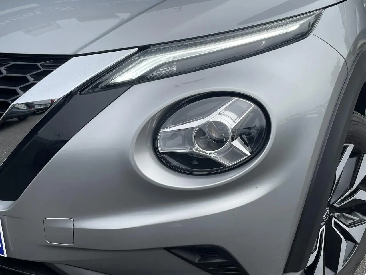 Gros plan sur le phare avant droit et la calandre d’un Nissan Juke 2024 gris silver avec jante alliage visible.