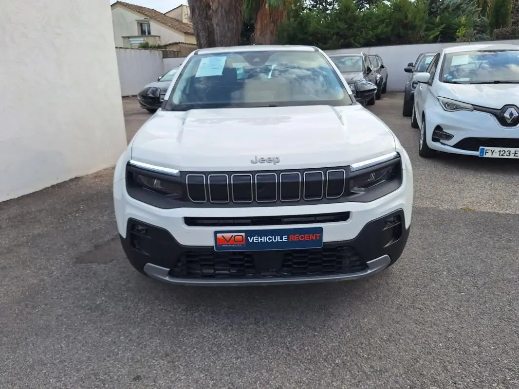 Vue frontale d'un Jeep Avenger blanc avec calandre noire et phares LED, stationné parmi d'autres véhicules.