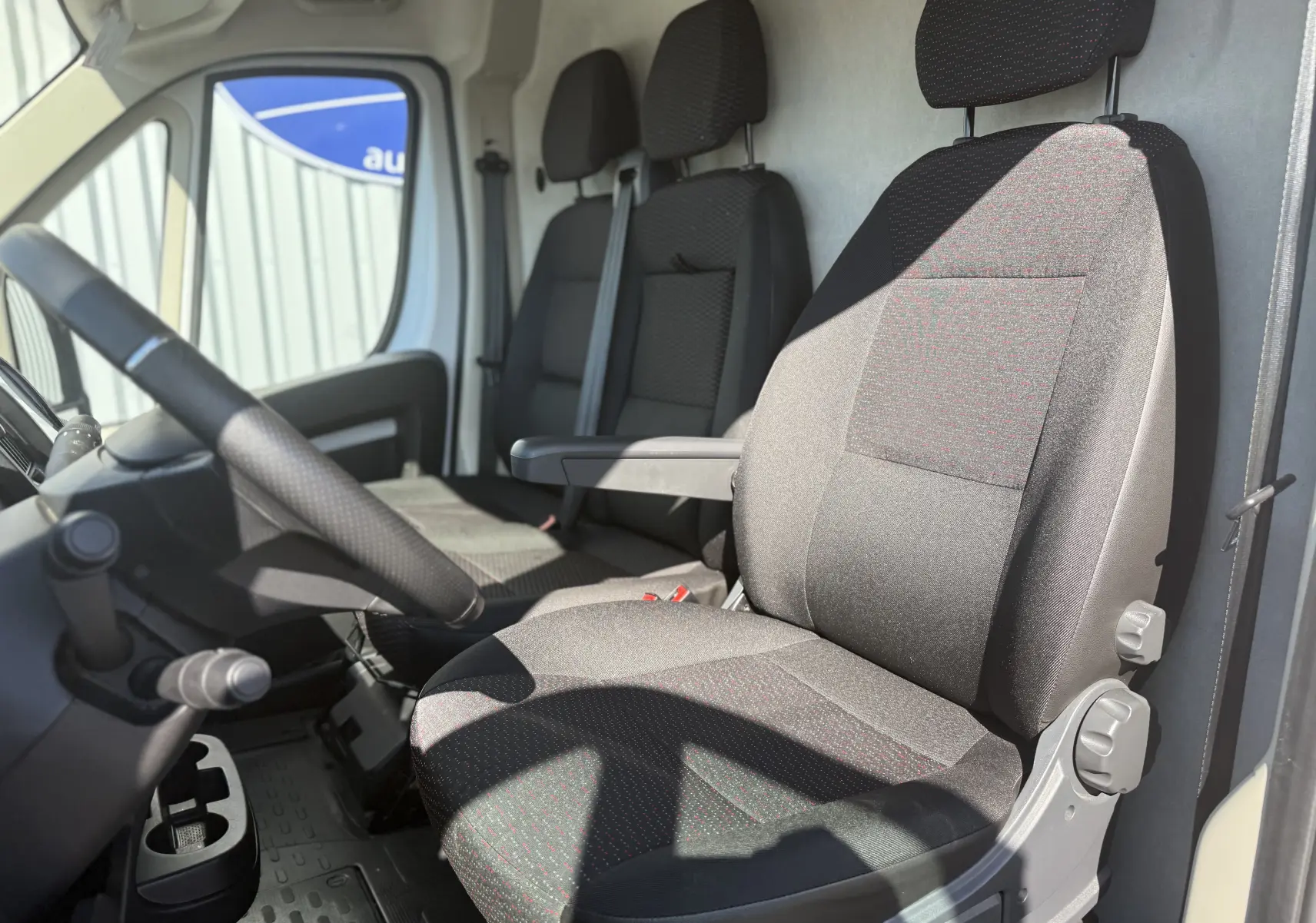 Vue intérieure côté conducteur de l’Opel Movano Fourgon blanc, montrant les sièges en tissu noir avec accoudoir.