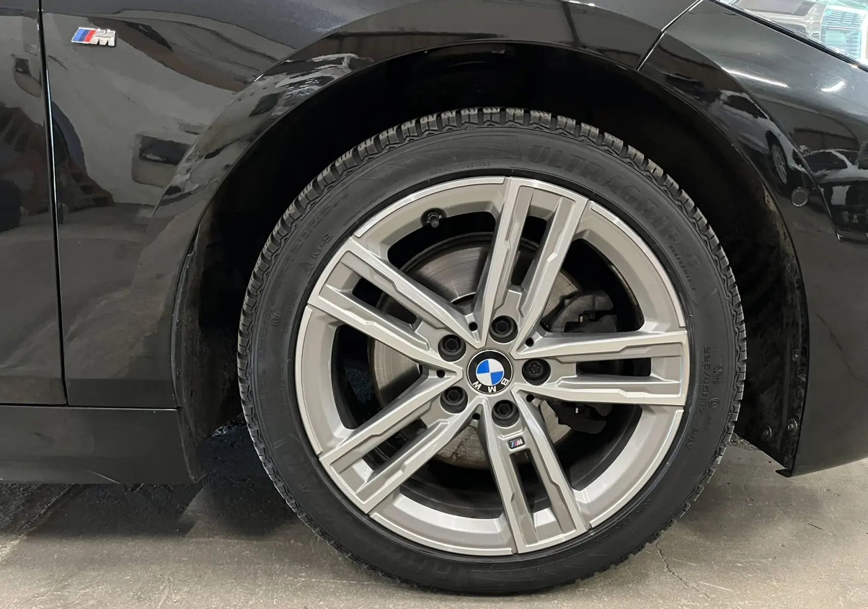 Gros plan sur la roue avant droite en alliage argenté avec logo BMW M sur une BMW Série 1 noire métallisée.