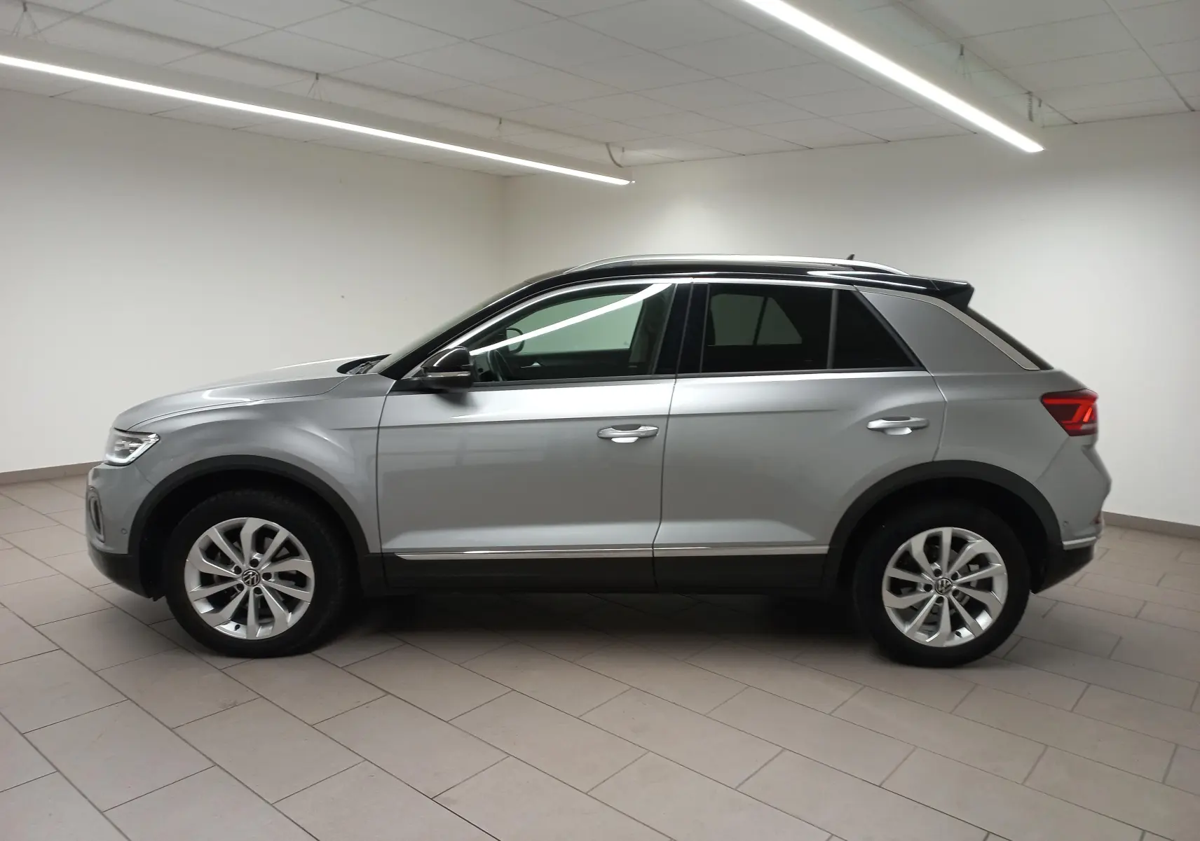 Profil côté gauche du Volkswagen T-Roc 2023 gris pyrite avec toit noir et vitres arrière surteintées en intérieur neutre.