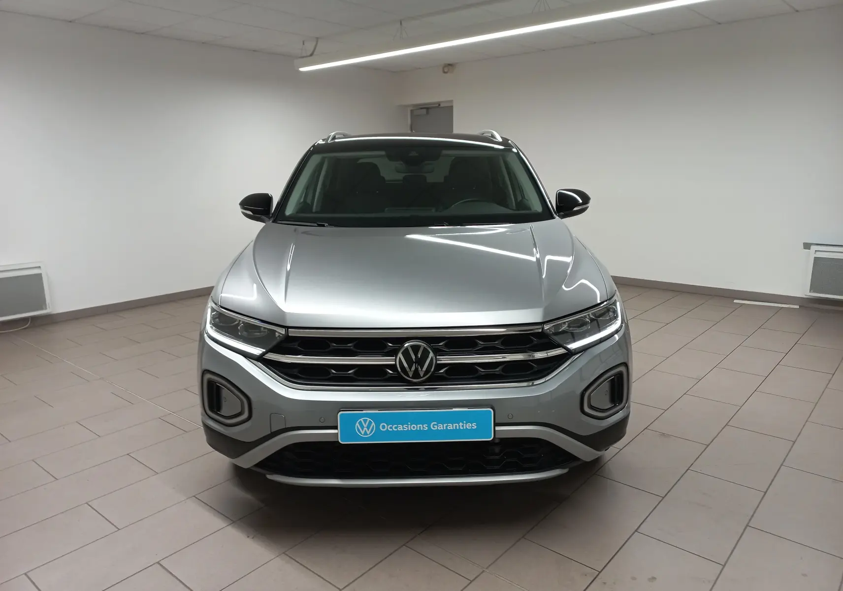 Volkswagen T-Roc gris pyrite vu de face dans un intérieur, avec toit noir et calandre chromée.