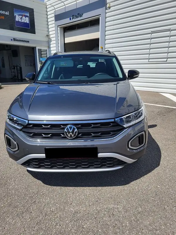 Vue frontale d'un Volkswagen T-Roc gris Indium 2025 garé devant un atelier, avec calandre et phares LED visibles.