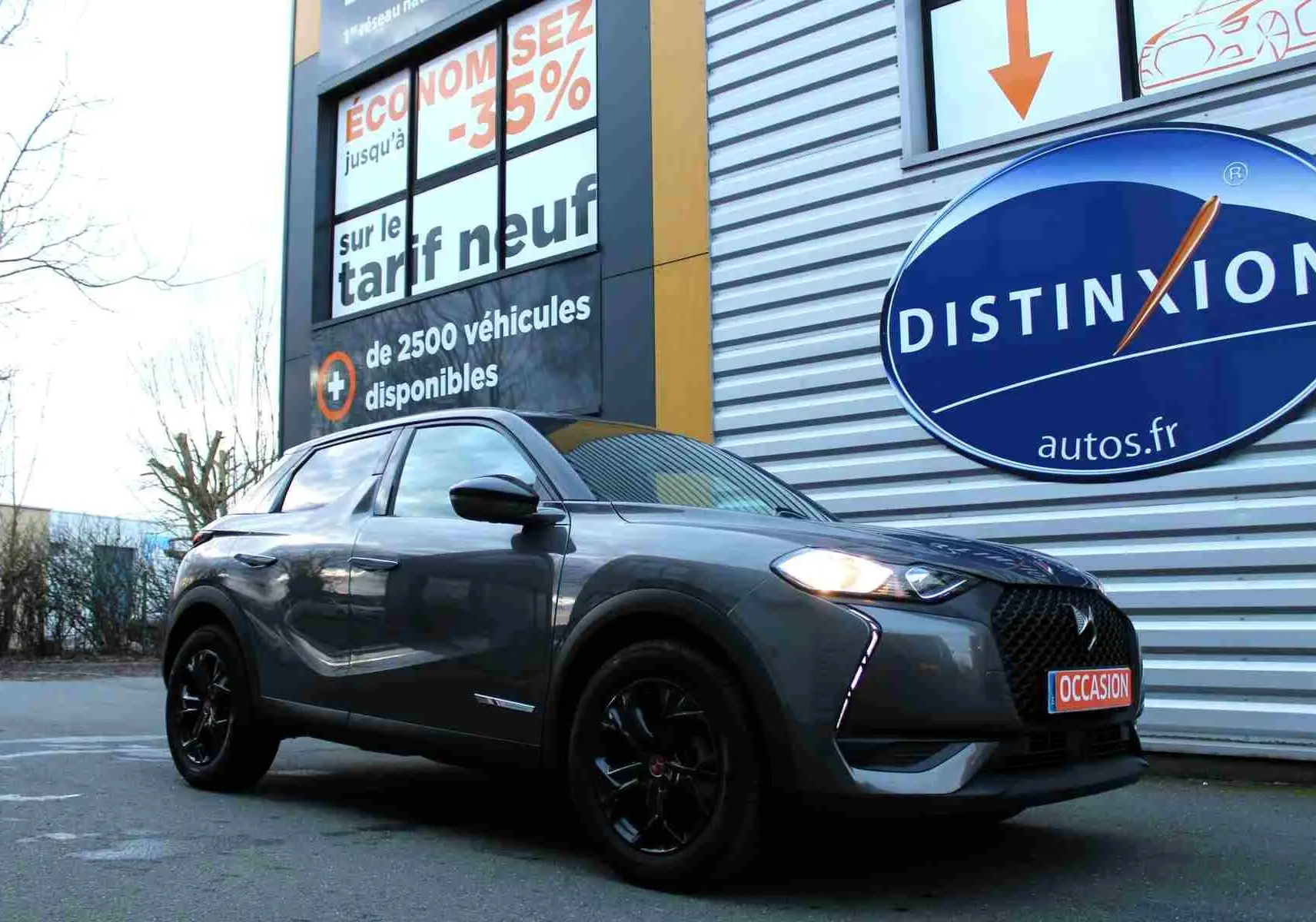 DS3 Crossback gris platinium métal en 3/4 avant droit, avec jantes noires et phares allumés, devant un panneau publicitaire.