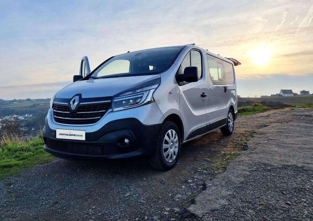 Renault Trafic Fourgon gris platine en 3/4 avant droit, porte latérale ouverte, sur chemin en extérieur au coucher du soleil.
