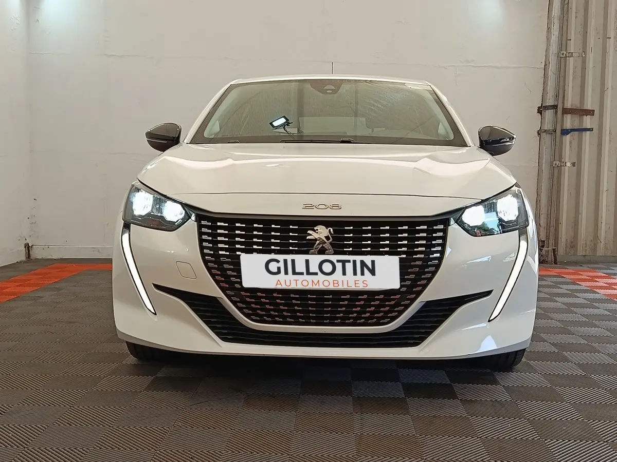 Vue frontale d'une Peugeot 208 blanche 2023 avec phares LED allumés et calandre noire distinctive.