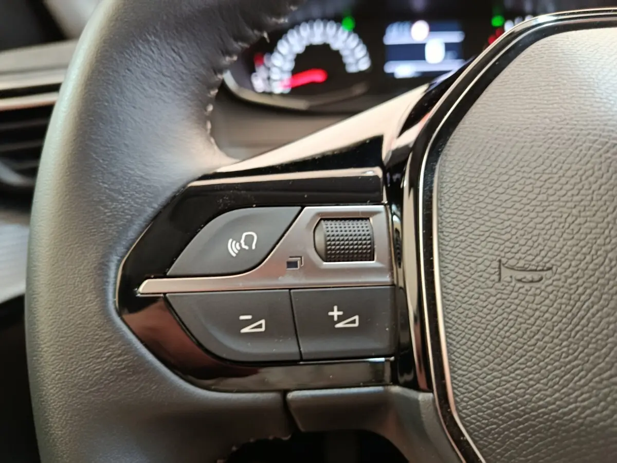 Gros plan sur les commandes gauche du volant cuir noir d’une Peugeot 208 blanche, avec tableau de bord flou en arrière-plan.