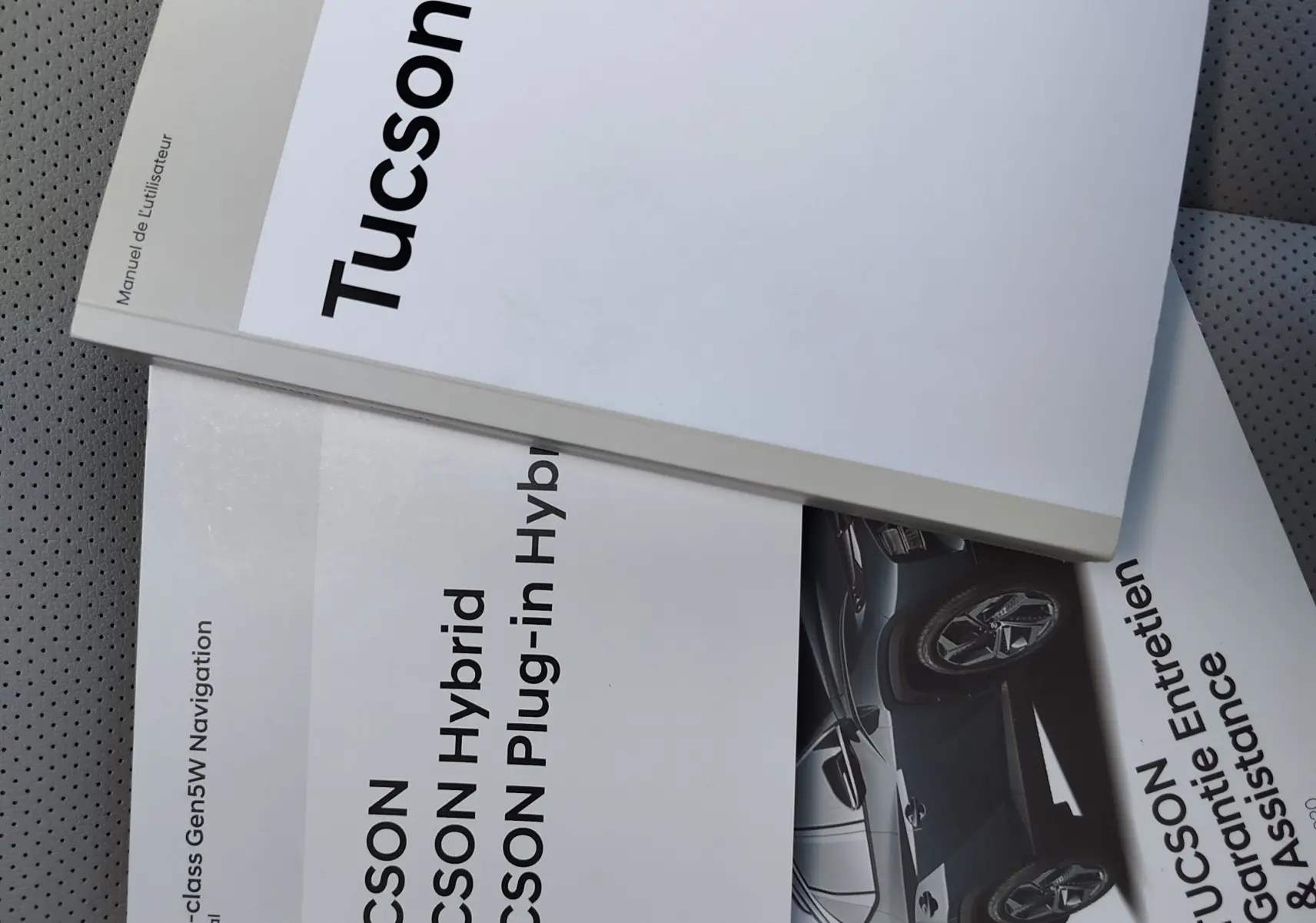 Manuels d'utilisation Hyundai Tucson posés en angle sur un siège perforé gris clair, avec image partielle du SUV bleu métallisé.
