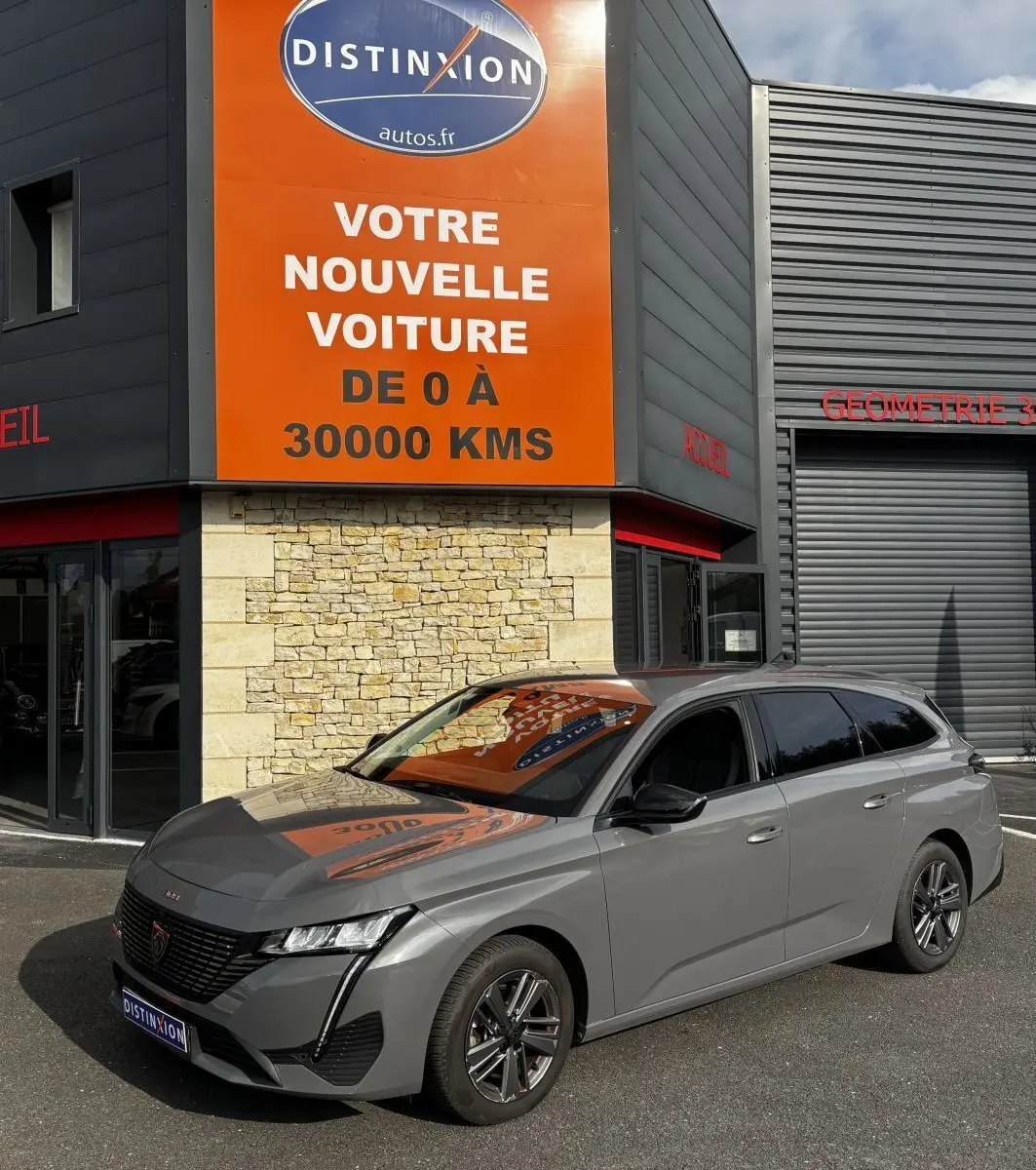 Peugeot 308 PureTech 130 S&S Allure gris clair vue 3/4 avant droit devant un bâtiment avec panneau orange.