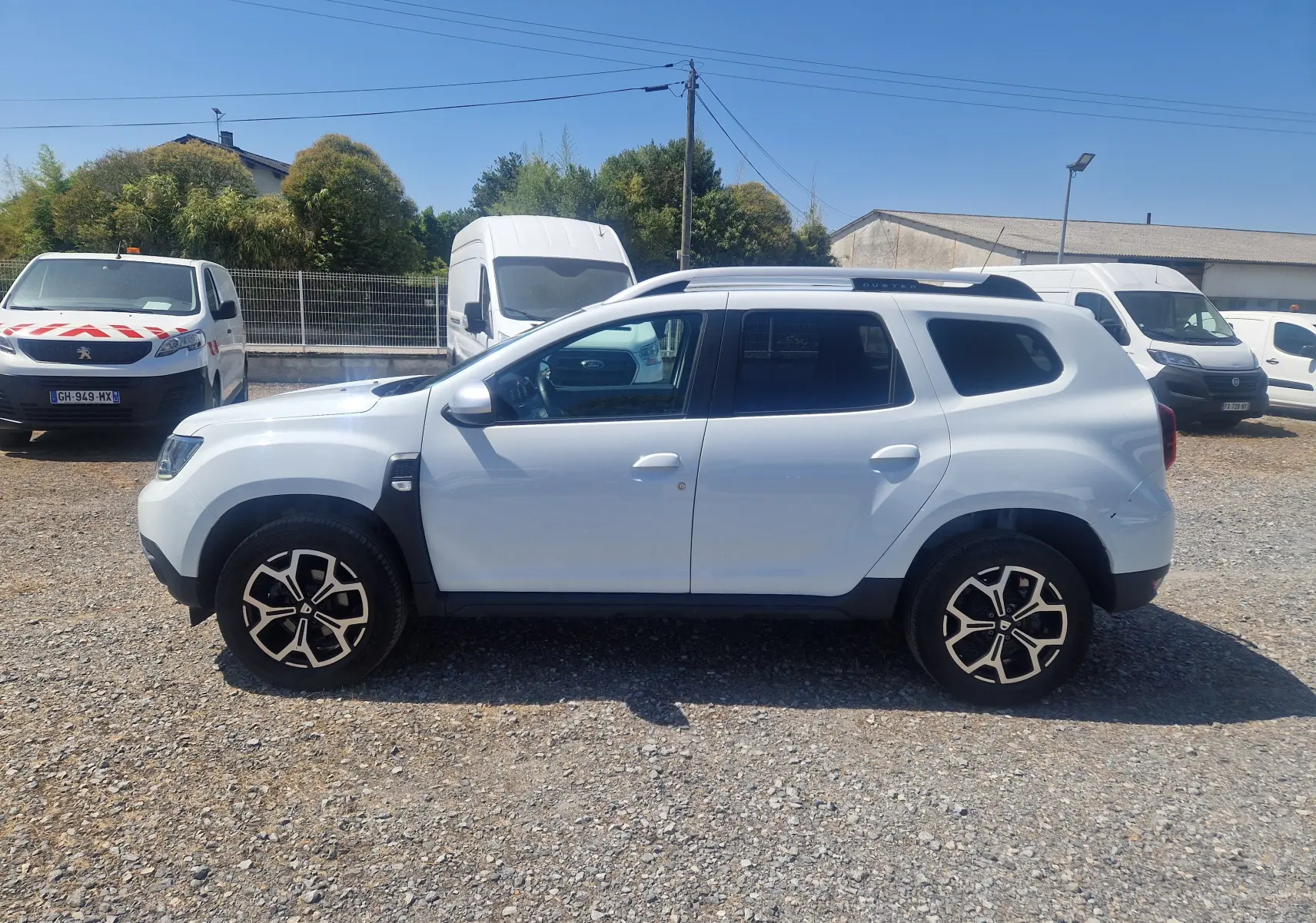 Profil droit d'un Dacia Duster blanc 2021 avec jantes alliage noires et toit noir, stationné sur un sol gravillonné.