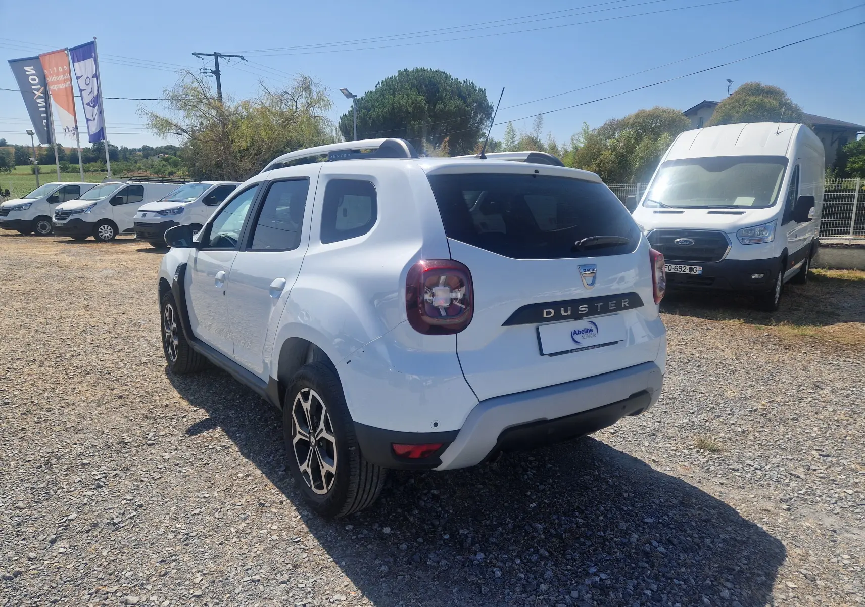 Vue 3/4 arrière droite d’un Dacia Duster blanc 2021 avec jantes alliage noires et toit ouvrant, stationné sur un terrain gravelé.