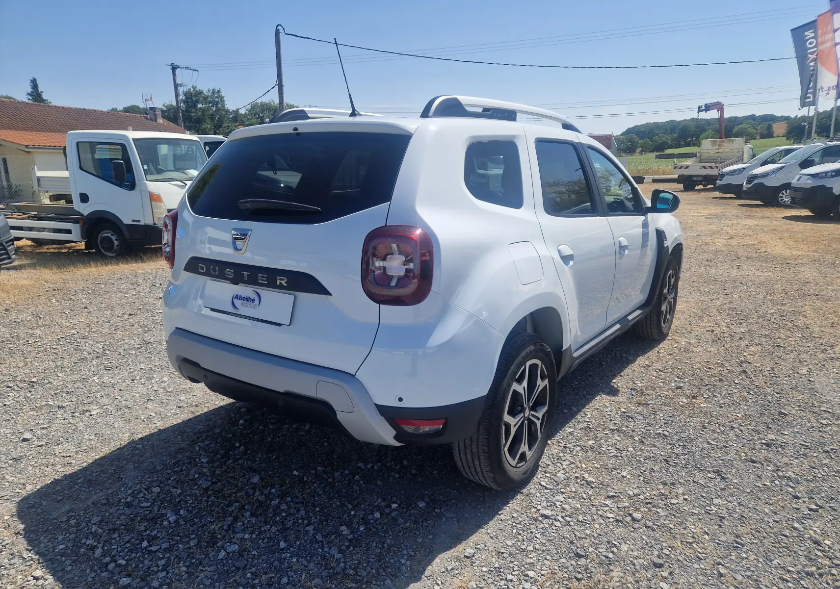 Vue 3/4 arrière droite du Dacia Duster blanc 2021 avec jantes alliage noires et barres de toit visibles sur un terrain gravelé.