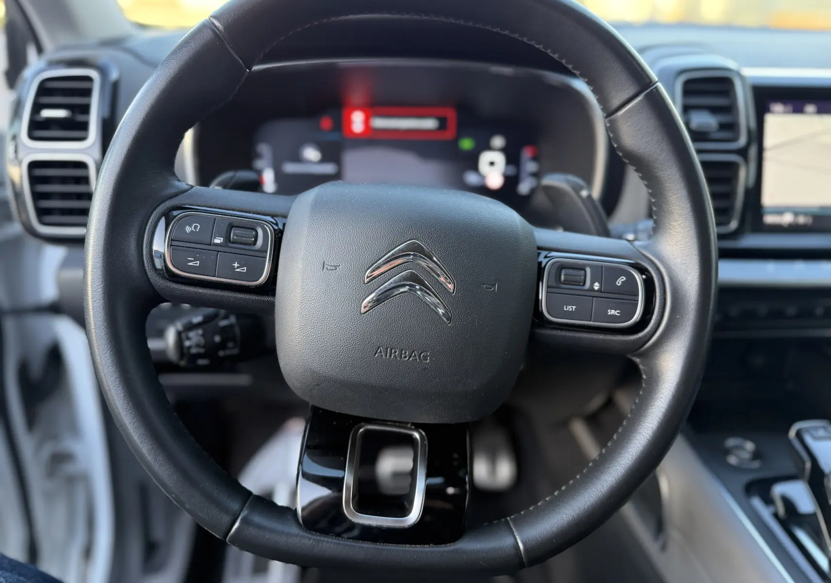 Vue rapprochée du volant cuir noir du Citroën C5 Aircross 2021, avec commandes intégrées et tableau de bord numérique flou.