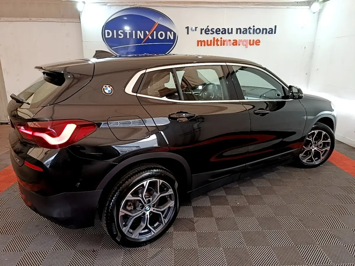 Vue 3/4 arrière droite d’un BMW X2 sDrive18i Lounge noir brillant avec jantes alliage et feux arrière LED allumés.