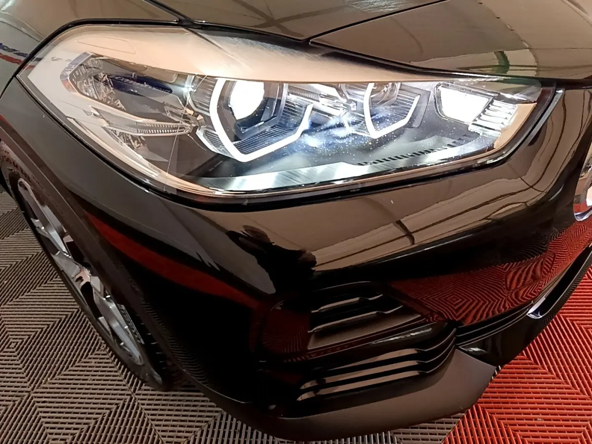 Gros plan sur le phare avant droit allumé d’un BMW X2 sDrive18i Lounge noir, avec détails du pare-chocs et jante visible.