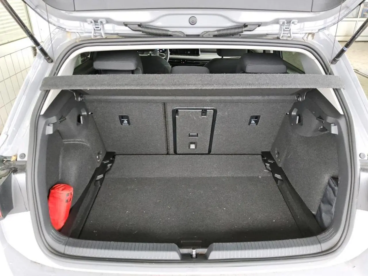 Coffre ouvert vu de l'arrière d'une Volkswagen Golf 8 gris lunaire, avec cache-bagages et trousse de secours rouge.