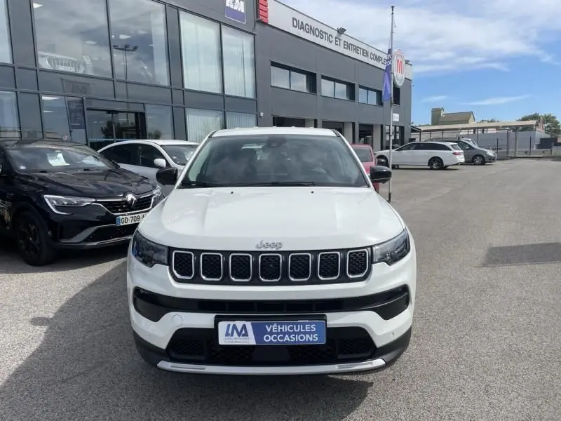 Vue frontale d’un Jeep Compass blanc 2024 avec calandre à sept fentes et plaque « Véhicules Occasions » devant un garage.
