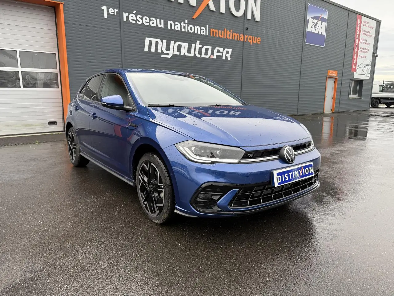 Volkswagen Polo 1.0 TSI R-Line bleu récif métallisé vue 3/4 avant droit sur sol mouillé devant un bâtiment.