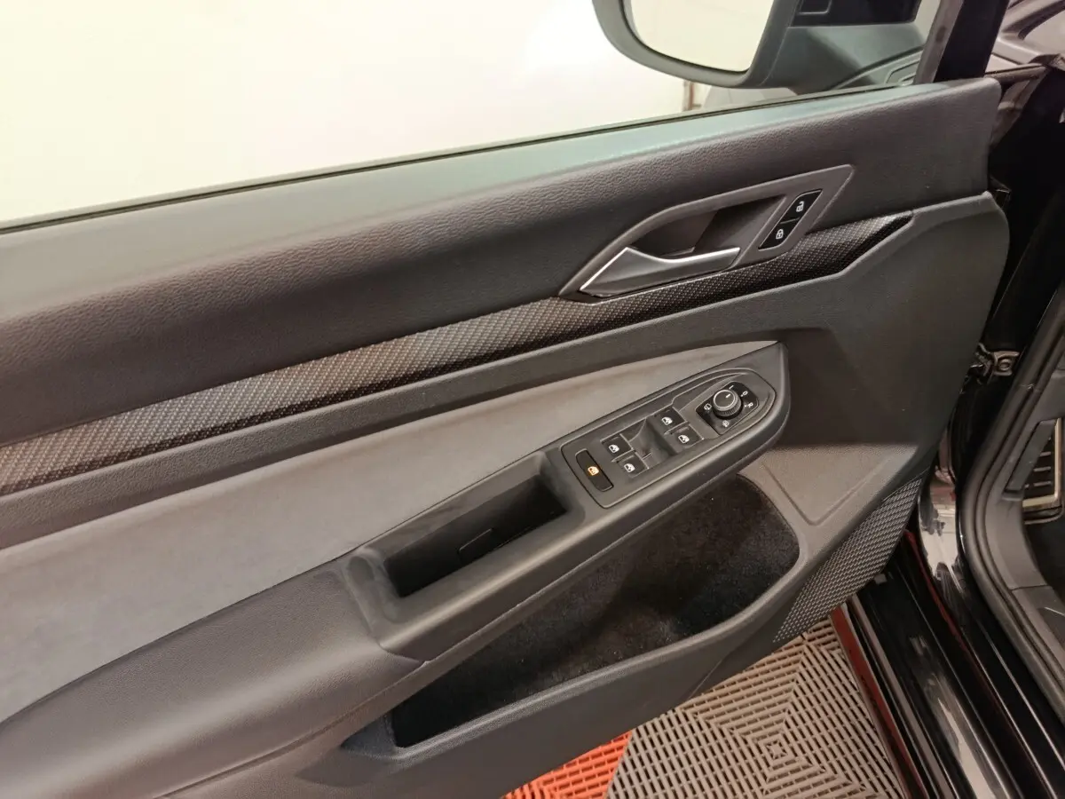 Vue rapprochée de la porte avant gauche noire d’une Volkswagen Golf 1.5 eTSI R-Line, avec commandes électriques et insert carbone.